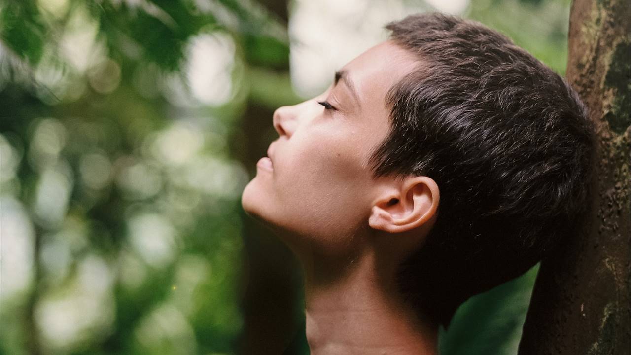 Mujer con los ojos cerrados apoyada en un árbol del bosque